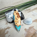 Handgefertigter, länglicher Surfbrett-Anhänger aus naturbelassenem Maserholz und transparentem Harz in Lagunen-Türkis, an schwarzem Lederband mit Knoten – maritimer Unikatschmuck für Damen und Herren, dekorativ mit Stein und Muschel inszeniert.