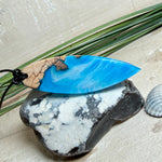 Handgefertigter Surfbrett-Anhänger aus warm gemasertem Olivenholz und Epoxidharz in Blau und Weiß, das an Meereswellen erinnert. Längliche, vorne polierte Form, ca. 6,2 × 2 cm, an verstellbarem schwarzem Lederband mit Knoten. Maritimer Unikatschmuck für Damen und Herren, dekorativ mit Stein und Muschel arrangiert.