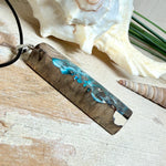 Rechteckiger Stabanhänger aus Maserholz und glasklarem Epoxidharz in tiefen Blau- und Türkistönen mit organischer Lagunen/Riff-Optik. Hochglanzpoliert, ca. 5,3 × 1,2 cm. Der Anhänger hängt an einer schwarzen Lederkette mit Edelstahl-Karabiner und 5-cm-Verlängerung (45 + 5 cm); die Anhängerschlaufe ist ebenfalls aus Edelstahl. Handgefertigtes Unikat, Lieferung in edler Schmuckbox – maritimer Look.