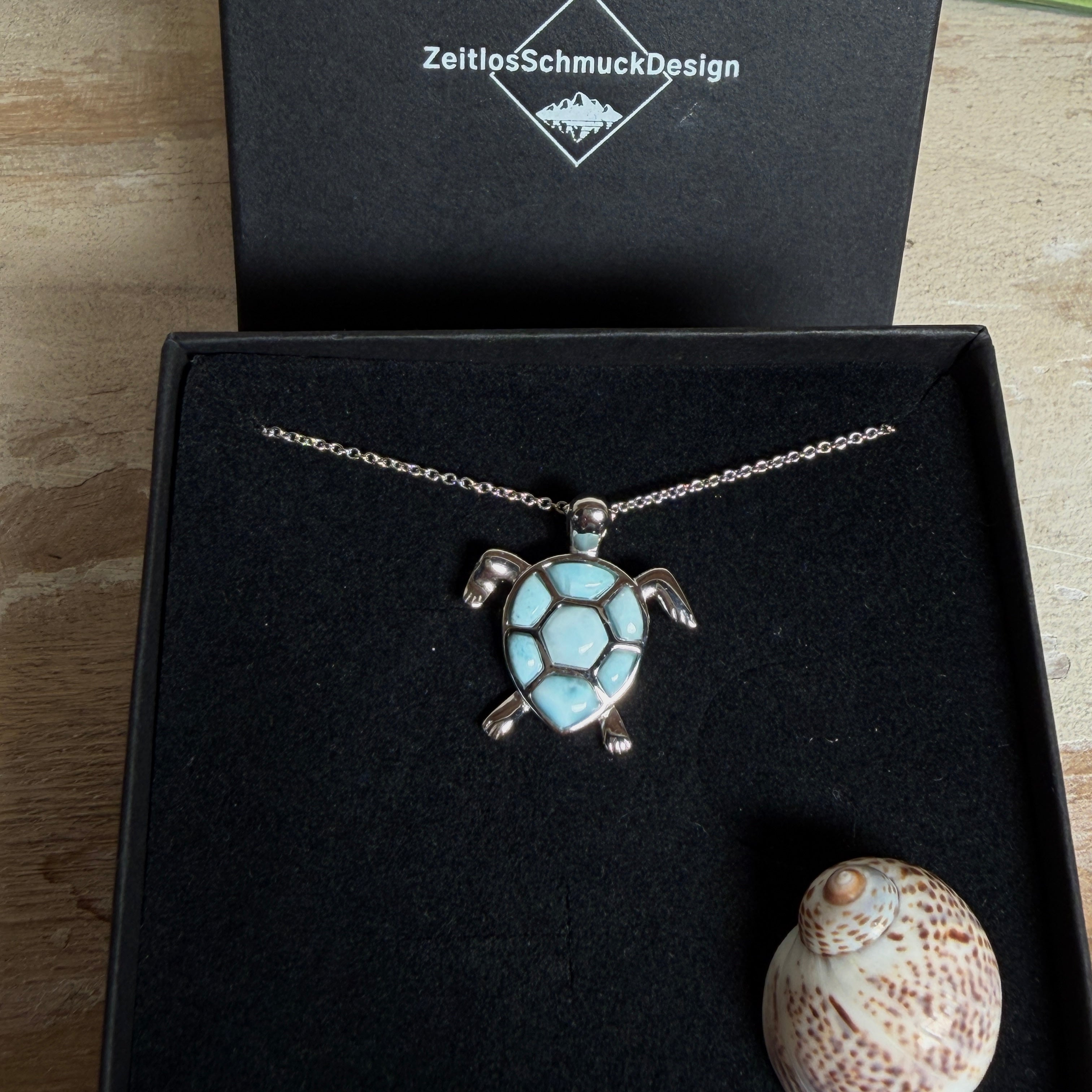 Feine Kette aus rhodiniertem 925 Silber mit Wasserschildkröten-Anhänger. Der ca. 25 x 18 mm große Anhänger zeigt eine Schildkröte, deren Panzer aus mehreren hellblauen Larimar-Inlays besteht. Seltener, kostbarer Larimar mit typischen wolkigen Strukturen, an einer ca. 45 cm langen, 1 mm starken, auf Platin rhodinierten Silberkette. Fotografiert in schwarzer Schmuckbox und vor maritimer Dekoration mit Muscheln.