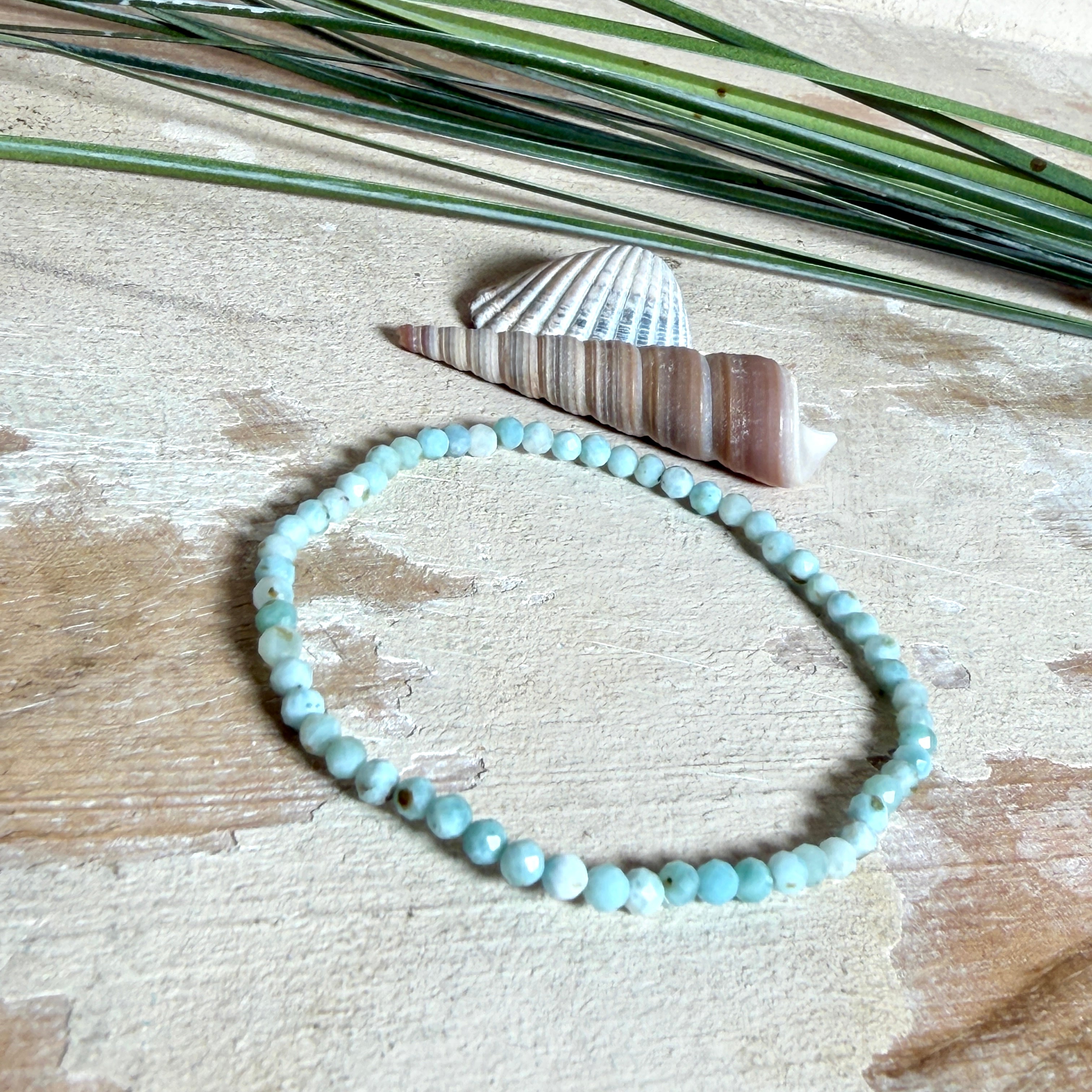 Zartes Larimar-Armband „Lagunenlicht“ mit facettierten 3 mm Perlen aus seltenem Atlantisstein auf Elastikfaden. Die kleinen Larimarsteine zeigen sanfte Blau- und Weißtöne wie Lagunenwasser. Innenumfang ca. 19 cm. Fotografiert auf Muscheln, einem dekorativen Stein und in einer schwarzen Schmuckbox von ZeitlosSchmuckDesign im maritimen Stil.
