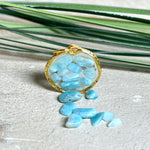 Runder Anhänger „Golden Atlantis Dream“ mit kleinen Larimar-Edelsteinchen in klarem Resin, gefasst in einer 24K vergoldeten Zinklegierung in Shiny Gold. Die Larimarsteine zeigen sanfte Blau- und Weißtöne wie karibische Wellen. Durchmesser ca. 22 mm, handgefertigtes Unikat, fotografiert mit Larimarsteinchen, Muscheln und einer 2-Euro-Münze zur Größenorientierung auf maritimem Untergrund.