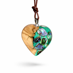 Herzförmiger Anhänger aus Olivenholz mit aquamarinfarbenem Harz und eingelegten Paua-Muschelstücken (Abalone), irisierender Blau-Grün-Schimmer. Der Anhänger hängt an einem braunen, verstellbaren Lederband mit Schiebeknoten; Aufhängung aus Edelstahl. Produktfoto vor maritimer Deko/Abalone-Schale.