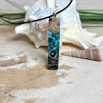 Rechteckiger Stabanhänger aus Maserholz und glasklarem Epoxidharz in tiefen Blau- und Türkistönen mit organischer Lagunen/Riff-Optik. Hochglanzpoliert, ca. 5,3 × 1,2 cm. Der Anhänger hängt an einer schwarzen Lederkette mit Edelstahl-Karabiner und 5-cm-Verlängerung (45 + 5 cm); die Anhängerschlaufe ist ebenfalls aus Edelstahl. Handgefertigtes Unikat, Lieferung in edler Schmuckbox – maritimer Look.