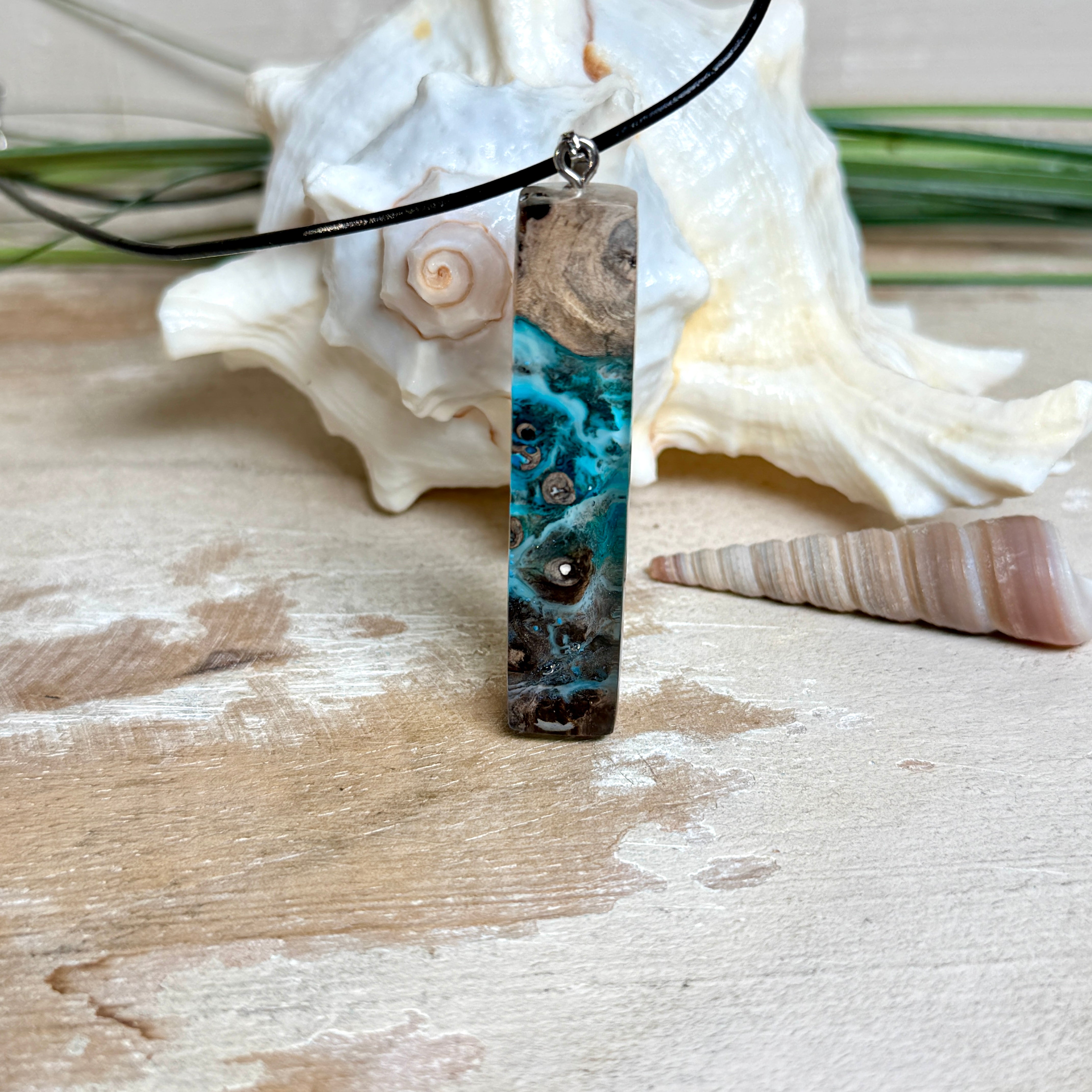 Rechteckiger Stabanhänger aus Maserholz und glasklarem Epoxidharz in tiefen Blau- und Türkistönen mit organischer Lagunen/Riff-Optik. Hochglanzpoliert, ca. 5,3 × 1,2 cm. Der Anhänger hängt an einer schwarzen Lederkette mit Edelstahl-Karabiner und 5-cm-Verlängerung (45 + 5 cm); die Anhängerschlaufe ist ebenfalls aus Edelstahl. Handgefertigtes Unikat, Lieferung in edler Schmuckbox – maritimer Look.