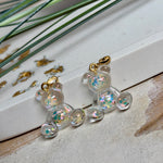 Kleiner Bärchen-Anhänger aus transparentem Harz mit irisierenden Flakes in Regenbogen-Schimmer, goldfarbene Anhängerschlaufe aus Edelstahl. Größe ca. 2,2 × 2,1 cm. Handgefertigtes Unikat; Flakes-Anordnung variiert, vereinzelt kleine Luftbläschen möglich.