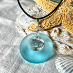 Handgefertigte Halskette mit Donut-Anhänger aus türkisfarbenem Seeglas, kombiniert mit einem filigranen Sonnen-Charm aus Edelstahl. Die Kette besteht aus einer schwarzen Lederschnur (1,5 mm) mit Karabiner und Verlängerungskette aus Edelstahl. Maritimes Schmuckstück für Meerliebhaberinnen und Sommerfreunde. Unikat mit stilvoller Ausstrahlung.
