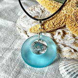 Handgefertigte Halskette mit Donut-Anhänger aus türkisfarbenem Seeglas, kombiniert mit einem filigranen Sonnen-Charm aus Edelstahl. Die Kette besteht aus einer schwarzen Lederschnur (1,5 mm) mit Karabiner und Verlängerungskette aus Edelstahl. Maritimes Schmuckstück für Meerliebhaberinnen und Sommerfreunde. Unikat mit stilvoller Ausstrahlung.