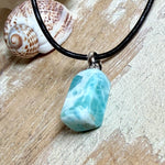 Larimar Freeform Anhänger an schwarzer Lederkette. Natürlicher, rechteckiger Larimar-Stein (ca. 1,5 × 1 cm) in Türkis-Blau mit weißen Strukturen, eingefasst in eine rhodinierte Silberöse. Beidseitig tragbar, an 45 cm Lederkette mit Edelstahl-Karabinerverschluss und 5 cm Verlängerung. Maritimes, zurückhaltend elegantes Schmuckstück mit der Optik karibischen Meerwassers.