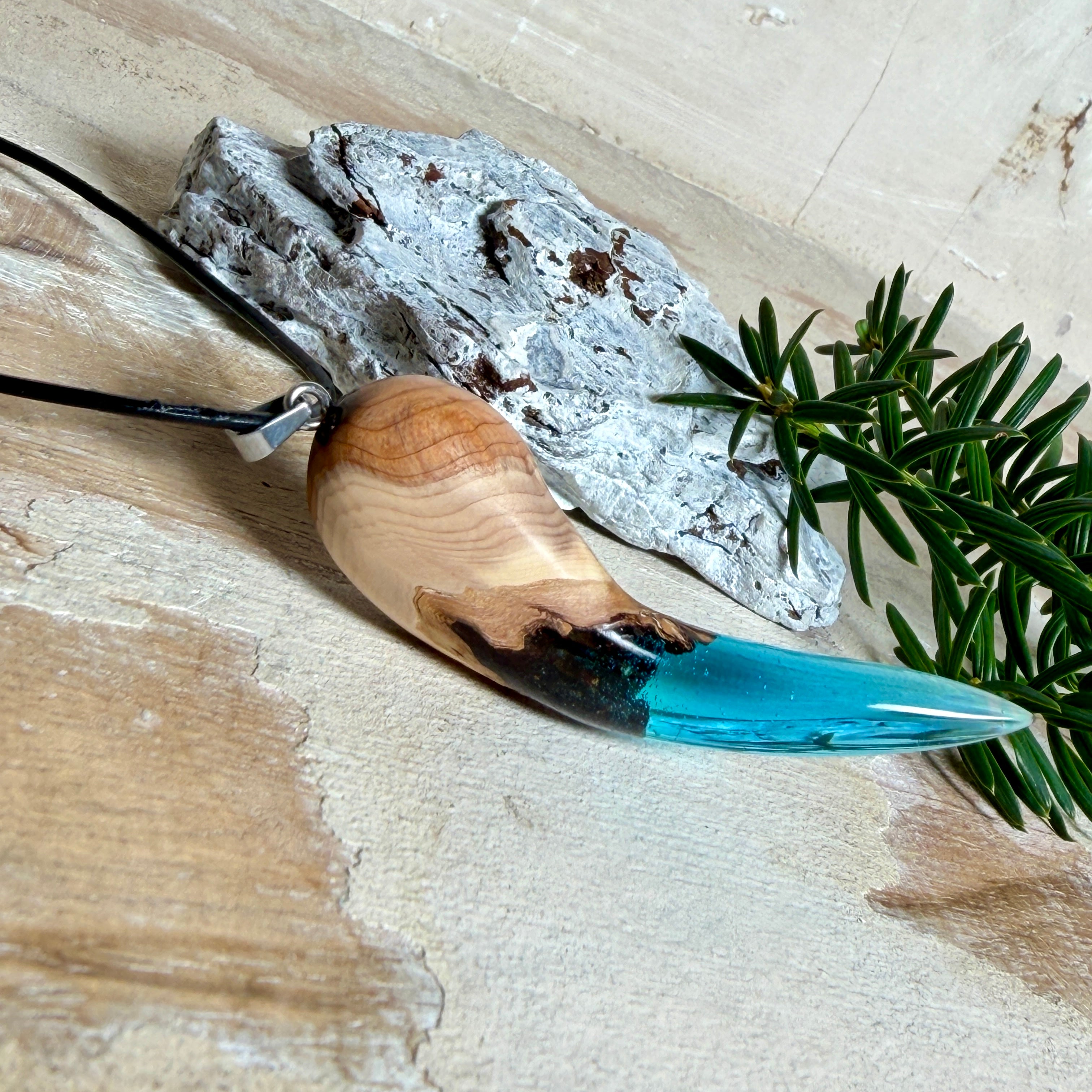 Handgefertigter Anhänger aus Edelholz und transparent-türkisblauem Epoxidharz, glatt poliert. Größe ca. 6,7 × 2,0 cm. Mit Edelstahlöse an schwarzer Lederkette (45 cm + 5 cm Edelstahl-Verlängerung, Karabiner). Maritimer, moderner Unisex-Schmuck mit natürlicher Holzmaserung; kleine Lufteinschlüsse sind charakteristisch.