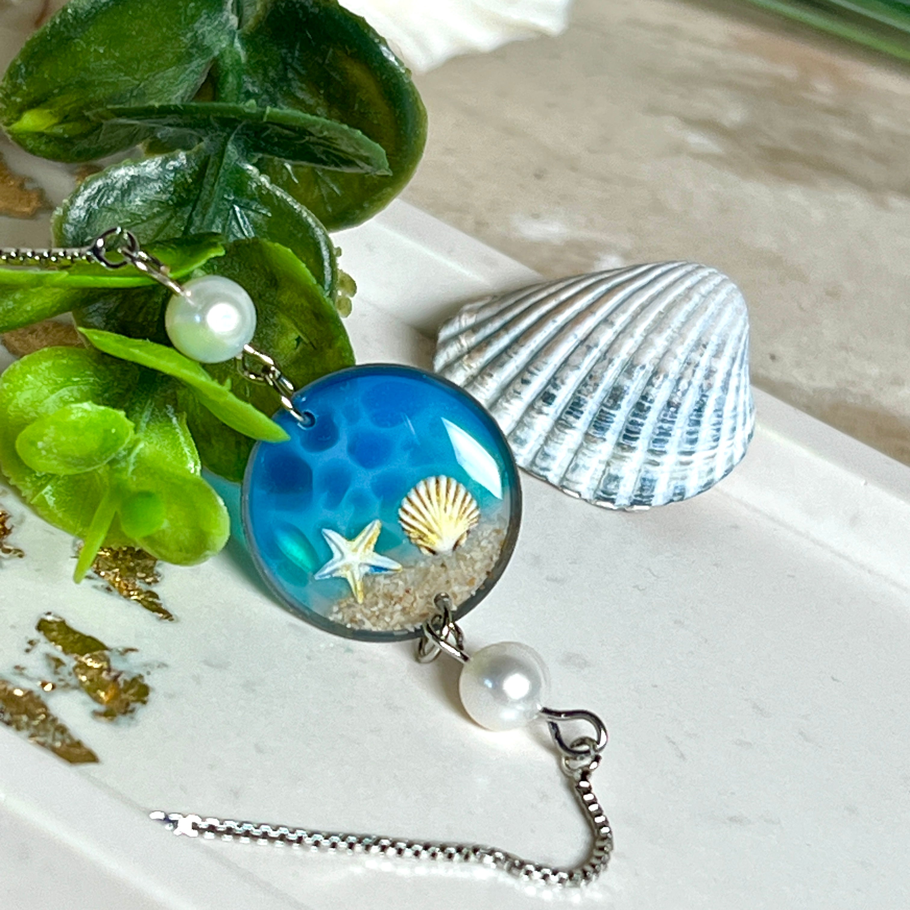 Elegantes Edelstahl-Armband mit rundem Resin-Anhänger in Meeresblau. Im Anhänger liegt echter Meersand als Ufersaum; darüber Sticker-Motive Seestern & Muschel. Zwei feine Süßwasserperlen verzieren das Armband. Verstellbarer Schiebeverschluss . Handgefertigtes Unikat – maritimer Schmuck, der an Wellenrauschen und Strandspaziergänge erinnert.