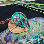 Herzförmiger Anhänger aus Olivenholz mit aquamarinfarbenem Harz und eingelegten Paua-Muschelstücken (Abalone), irisierender Blau-Grün-Schimmer. Der Anhänger hängt an einem braunen, verstellbaren Lederband mit Schiebeknoten; Aufhängung aus Edelstahl. Produktfoto vor maritimer Deko/Abalone-Schale.