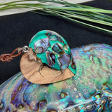 Herzförmiger Anhänger aus Olivenholz mit aquamarinfarbenem Harz und eingelegten Paua-Muschelstücken (Abalone), irisierender Blau-Grün-Schimmer. Der Anhänger hängt an einem braunen, verstellbaren Lederband mit Schiebeknoten; Aufhängung aus Edelstahl. Produktfoto vor maritimer Deko/Abalone-Schale.