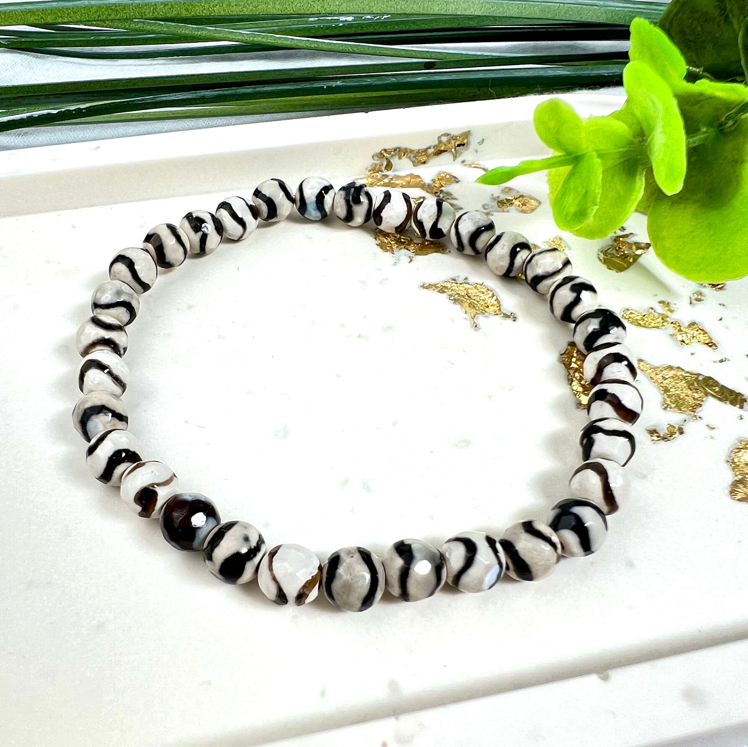 “Stilvolles Zebra-Achat-Armband mit schwarz-weißen Streifen auf hellem Untergrund, verziert mit goldenen Akzenten und grünen Blättern – ideal für Energiearbeit, spirituelle Balance und als modisches Accessoire.”