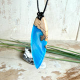 Handgefertigter Surfbrett-Anhänger aus warm gemasertem Olivenholz und Epoxidharz in Blau und Weiß, das an Meereswellen erinnert. Längliche, vorne polierte Form, ca. 6,2 × 2 cm, an verstellbarem schwarzem Lederband mit Knoten. Maritimer Unikatschmuck für Damen und Herren, dekorativ mit Stein und Muschel arrangiert.