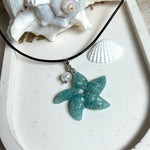Seestern-Anhänger aus transparentem Harz mit echtem Meersand ( leicht eingefärbt), in blautürkis Strand-Look, mit kleiner Süßwasserperle als Anhänger-Detail. Edelstahlöse, schwarze Lederkette mit Edelstahl-Karabiner und Verlängerung (45 cm + 5 cm). Anhänger ca. 3,5 × 3,5 cm. Handgefertigtes Unikat; vereinzelt kleine Luftbläschen möglich, typisch für Handarbeit.