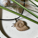 Handgefertigter Ammonit-Anhänger aus echtem Meersand & Harz mit filigraner Edelstahlöse und einer edlen Süßwasserperle. Maritimer Schmuck, inspiriert von der Kraft und Schönheit des Ozeans. Jedes Stück ein Unikat. Präsentiert auf einer Muschel, mit Lederkette.