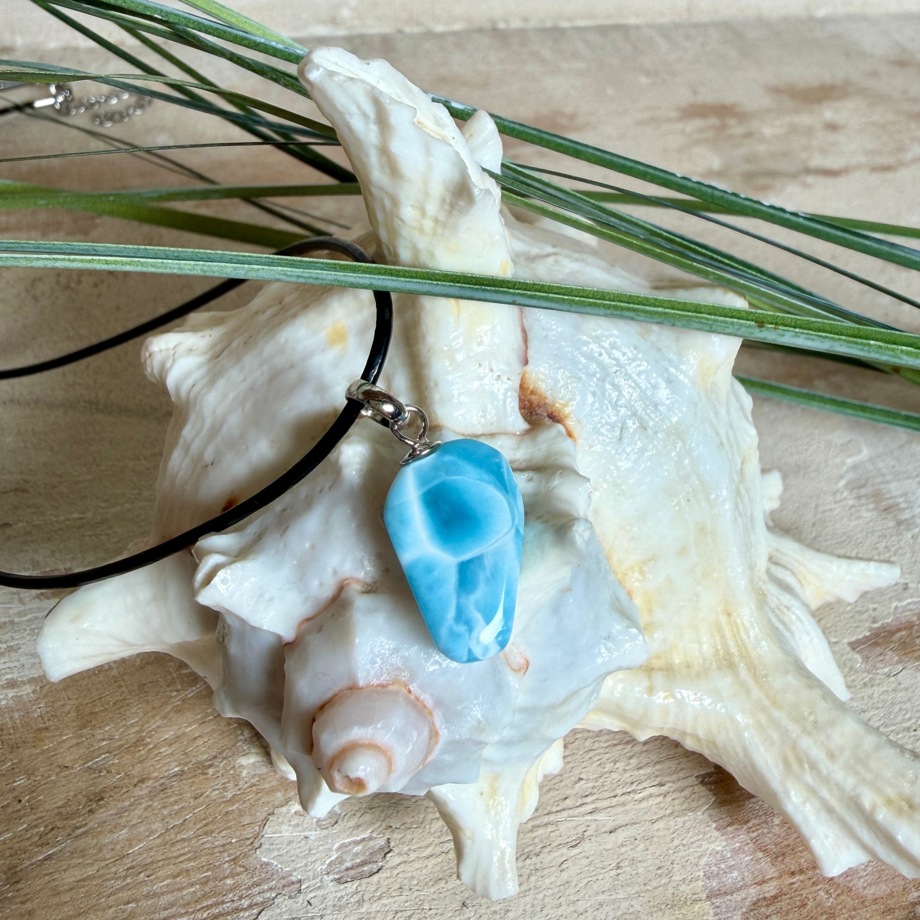 Larimar Freeform-Anhänger auf schwarzer Lederkette. Beidseitig tragbarer Trommelstein, ca. 2 x 1,8 cm groß, mit Silberöse. Der seltene, kostbare Larimar zeigt lebendige Blau- und Weißnuancen wie karibisches Lagunenwasser. Lederkette 45 cm mit Edelstahl-Karabiner und 5 cm Edelstahl-Verlängerung, fotografiert vor einer großen Muschel im maritimen Stil.