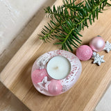 Rosa Teelichthalter aus klarem Epoxidharz, ca. 8 × 6,5 cm, Höhe ca. 4 cm. Im Harz sind kleine rosa Weihnachtskugeln, ein Stern, eine Schneeflocke und rosa Granulat eingegossen. Ovaler Halter für ein Teelicht, Unterseite mit vier runden Korkpads als Möbelschoner. Fotografiert auf Holz mit Tannenzweig und rosa Kugeln als weihnachtlicher Dekohintergrund.