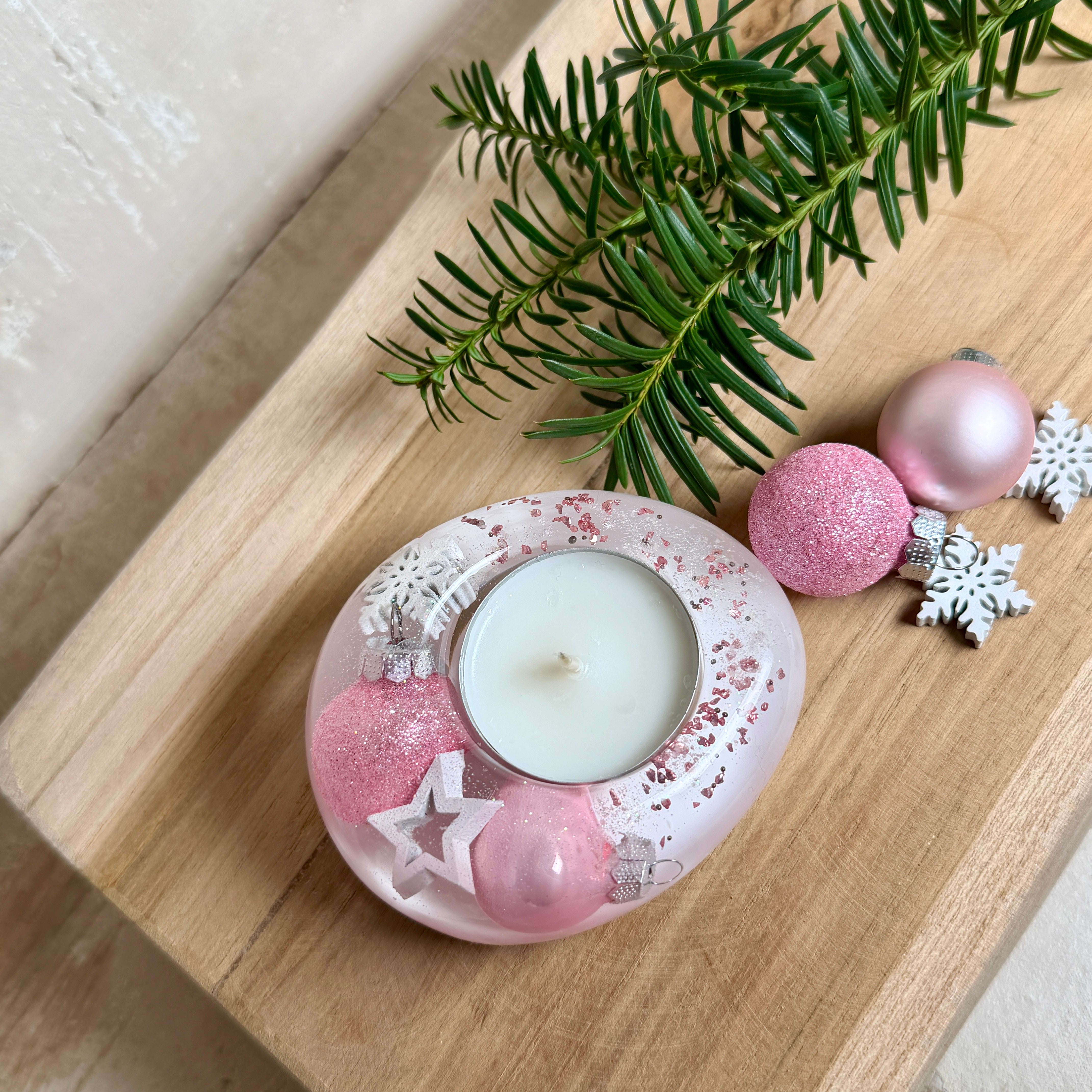 Rosa Teelichthalter aus klarem Epoxidharz, ca. 8 × 6,5 cm, Höhe ca. 4 cm. Im Harz sind kleine rosa Weihnachtskugeln, ein Stern, eine Schneeflocke und rosa Granulat eingegossen. Ovaler Halter für ein Teelicht, Unterseite mit vier runden Korkpads als Möbelschoner. Fotografiert auf Holz mit Tannenzweig und rosa Kugeln als weihnachtlicher Dekohintergrund.