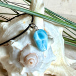Larimar Freeform-Anhänger auf schwarzer Lederkette. Beidseitig tragbarer Trommelstein, ca. 2 x 1,8 cm groß, mit Silberöse. Der seltene, kostbare Larimar zeigt lebendige Blau- und Weißnuancen wie karibisches Lagunenwasser. Lederkette 45 cm mit Edelstahl-Karabiner und 5 cm Edelstahl-Verlängerung, fotografiert vor einer großen Muschel im maritimen Stil.