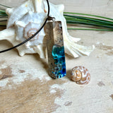 Rechteckiger Stabanhänger aus Maserholz und glasklarem Epoxidharz in tiefen Blau- und Türkistönen mit organischer Lagunen/Riff-Optik. Hochglanzpoliert, ca. 5,3 × 1,2 cm. Der Anhänger hängt an einer schwarzen Lederkette mit Edelstahl-Karabiner und 5-cm-Verlängerung (45 + 5 cm); die Anhängerschlaufe ist ebenfalls aus Edelstahl. Handgefertigtes Unikat, Lieferung in edler Schmuckbox – maritimer Look.