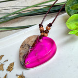 Handgefertigter Herzanhänger aus Olivenholz und leuchtend pinkem Epoxidharz mit eingeschlossenen Pusteblumensamen. Verstellbares braunes Lederband mit Holzperlen, Anhänger ca. 3,5 × 3,3 cm groß. Das Harz leuchtet dezent unter Schwarzlicht. Ein einzigartiges Unikat von ZeitlosSchmuckDesign, inspiriert von Kindheitsglück und Pusteblumen.