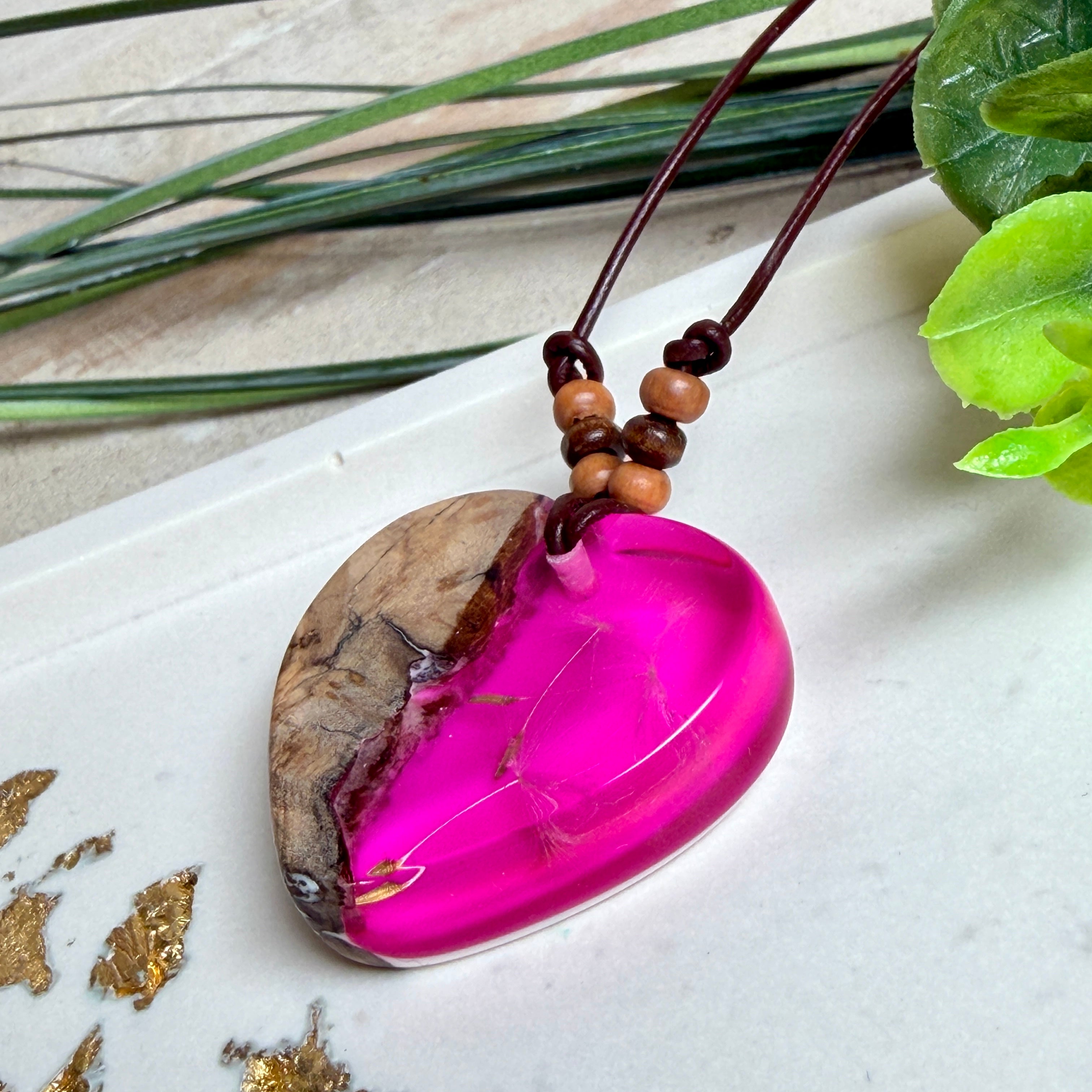 Handgefertigter Herzanhänger aus Olivenholz und leuchtend pinkem Epoxidharz mit eingeschlossenen Pusteblumensamen. Verstellbares braunes Lederband mit Holzperlen, Anhänger ca. 3,5 × 3,3 cm groß. Das Harz leuchtet dezent unter Schwarzlicht. Ein einzigartiges Unikat von ZeitlosSchmuckDesign, inspiriert von Kindheitsglück und Pusteblumen.