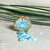 Runder Anhänger „Golden Atlantis Dream“ mit kleinen Larimar-Edelsteinchen in klarem Resin, gefasst in einer 24K vergoldeten Zinklegierung in Shiny Gold. Die Larimarsteine zeigen sanfte Blau- und Weißtöne wie karibische Wellen. Durchmesser ca. 22 mm, handgefertigtes Unikat, fotografiert mit Larimarsteinchen, Muscheln und einer 2-Euro-Münze zur Größenorientierung auf maritimem Untergrund.