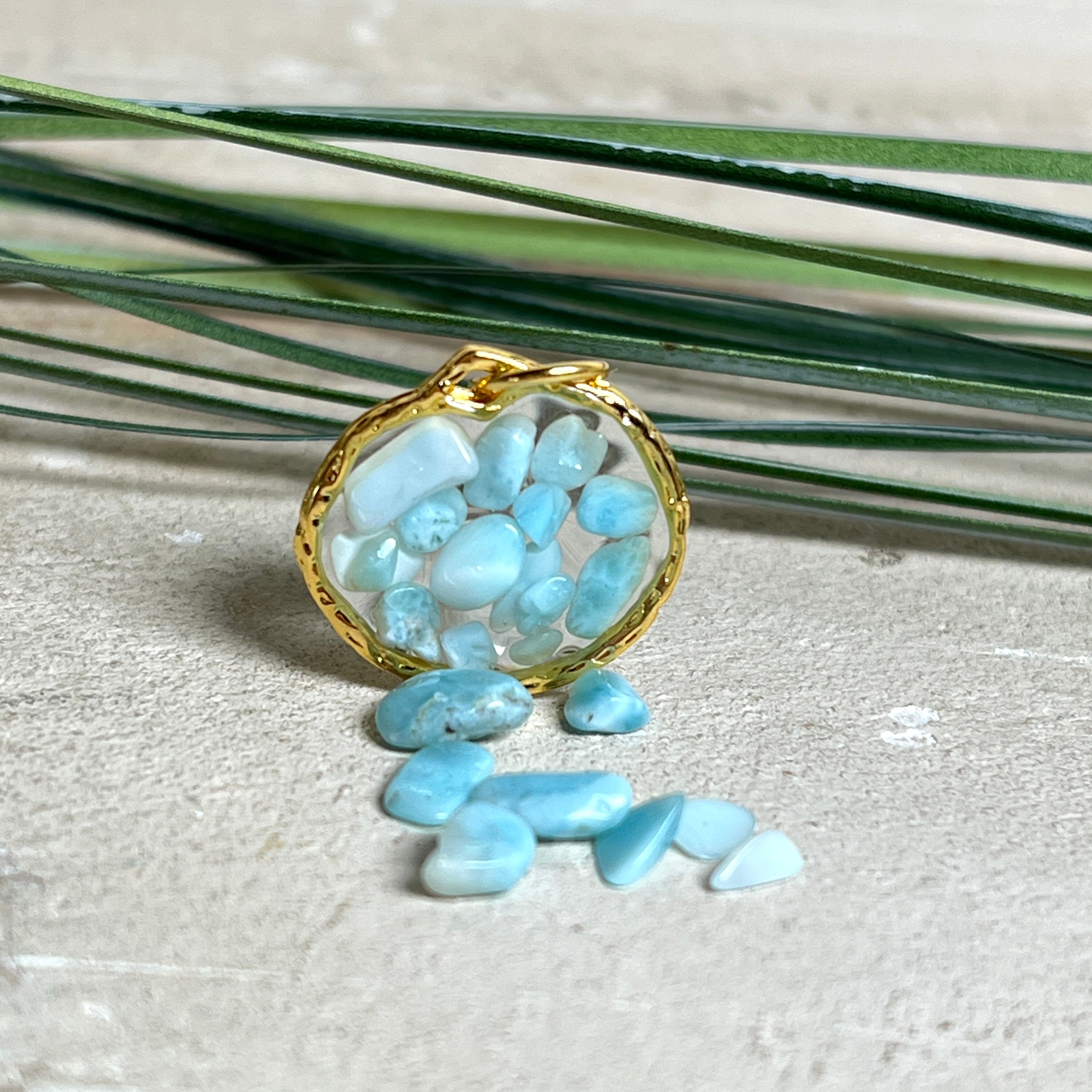 Runder Anhänger „Golden Atlantis Dream“ mit kleinen Larimar-Edelsteinchen in klarem Resin, gefasst in einer 24K vergoldeten Zinklegierung in Shiny Gold. Die Larimarsteine zeigen sanfte Blau- und Weißtöne wie karibische Wellen. Durchmesser ca. 22 mm, handgefertigtes Unikat, fotografiert mit Larimarsteinchen, Muscheln und einer 2-Euro-Münze zur Größenorientierung auf maritimem Untergrund.