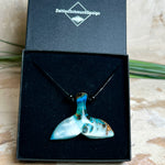 Handgefertigter Walflossen-Anhänger „Flossenklang“ aus Maserholz und türkis-blau-grau-weiß marmoriertem Epoxidharz an verstellbarem Lederband. Maritime Kette als Unikatschmuck für Meeresliebhaber und Wal-Fans, hochwertig poliert, präsentiert vor Muscheldeko und in Schmuckbox von ZeitlosSchmuckDesign.