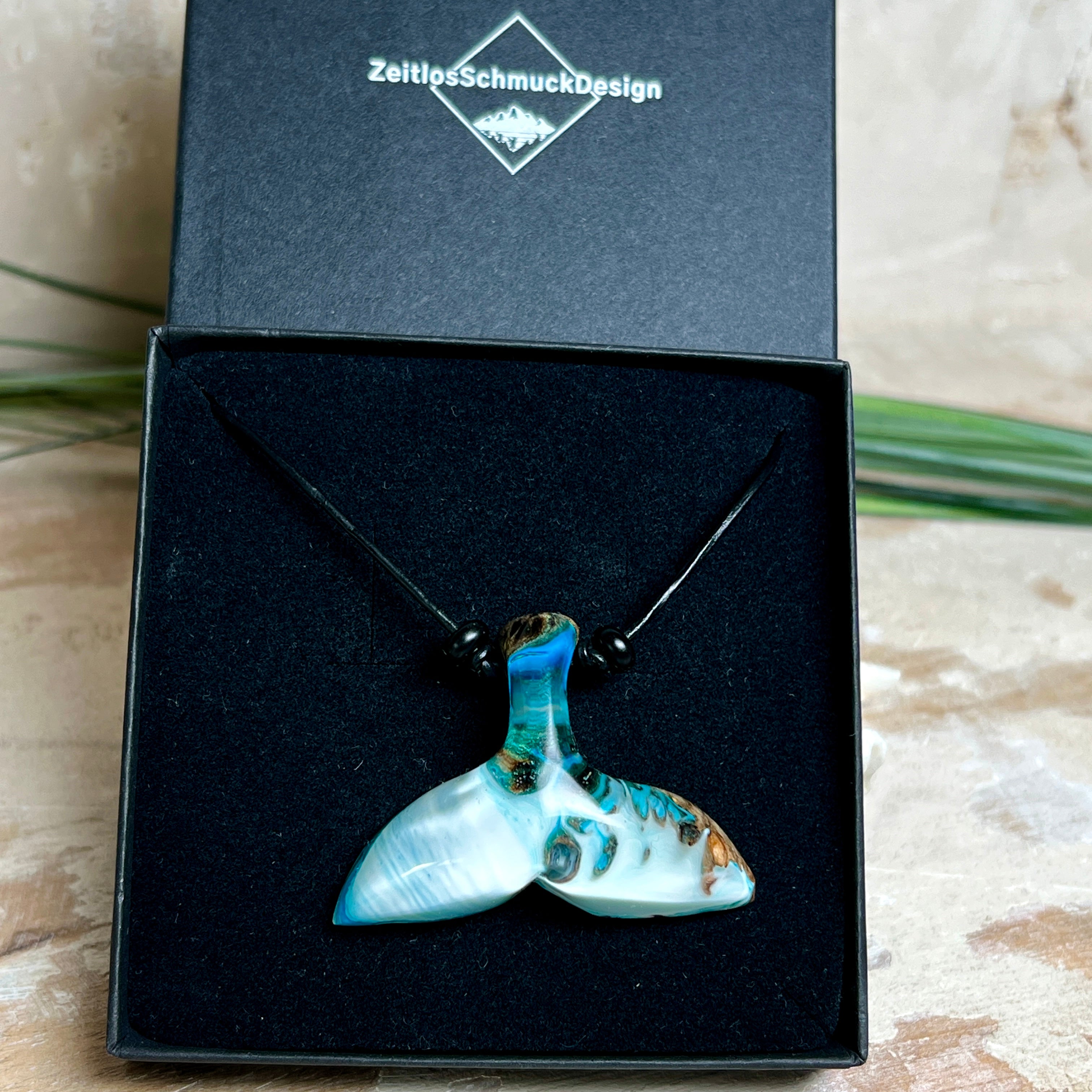 Handgefertigter Walflossen-Anhänger „Flossenklang“ aus Maserholz und türkis-blau-grau-weiß marmoriertem Epoxidharz an verstellbarem Lederband. Maritime Kette als Unikatschmuck für Meeresliebhaber und Wal-Fans, hochwertig poliert, präsentiert vor Muscheldeko und in Schmuckbox von ZeitlosSchmuckDesign.