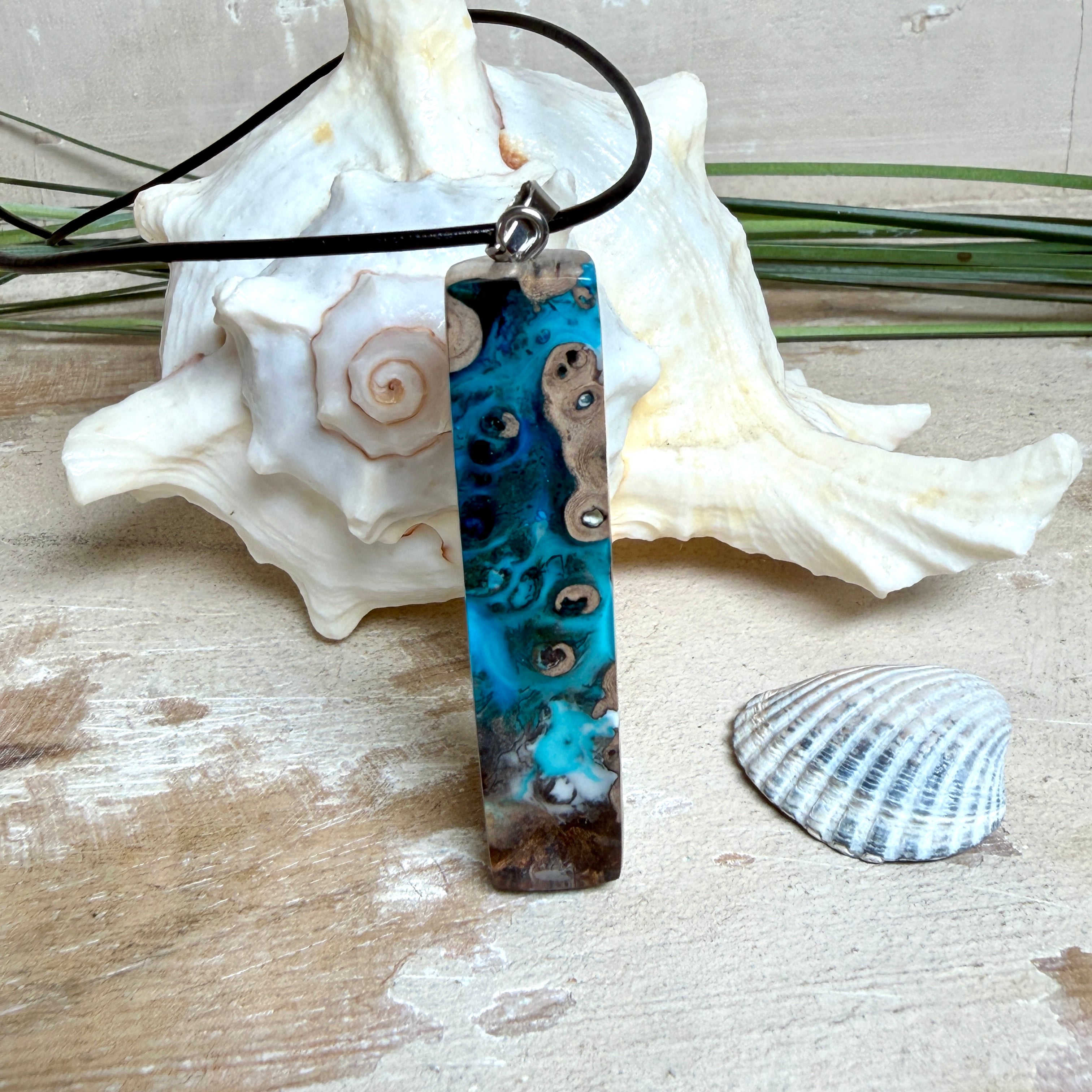 Rechteckiger Stabanhänger aus Maserholz und glasklarem Epoxidharz in tiefen Blau- und Türkistönen mit organischer Lagunen/Riff-Optik. Hochglanzpoliert, ca. 5,3 × 1,2 cm. Der Anhänger hängt an einer schwarzen Lederkette mit Edelstahl-Karabiner und 5-cm-Verlängerung (45 + 5 cm); die Anhängerschlaufe ist ebenfalls aus Edelstahl. Handgefertigtes Unikat, Lieferung in edler Schmuckbox – maritimer Look.