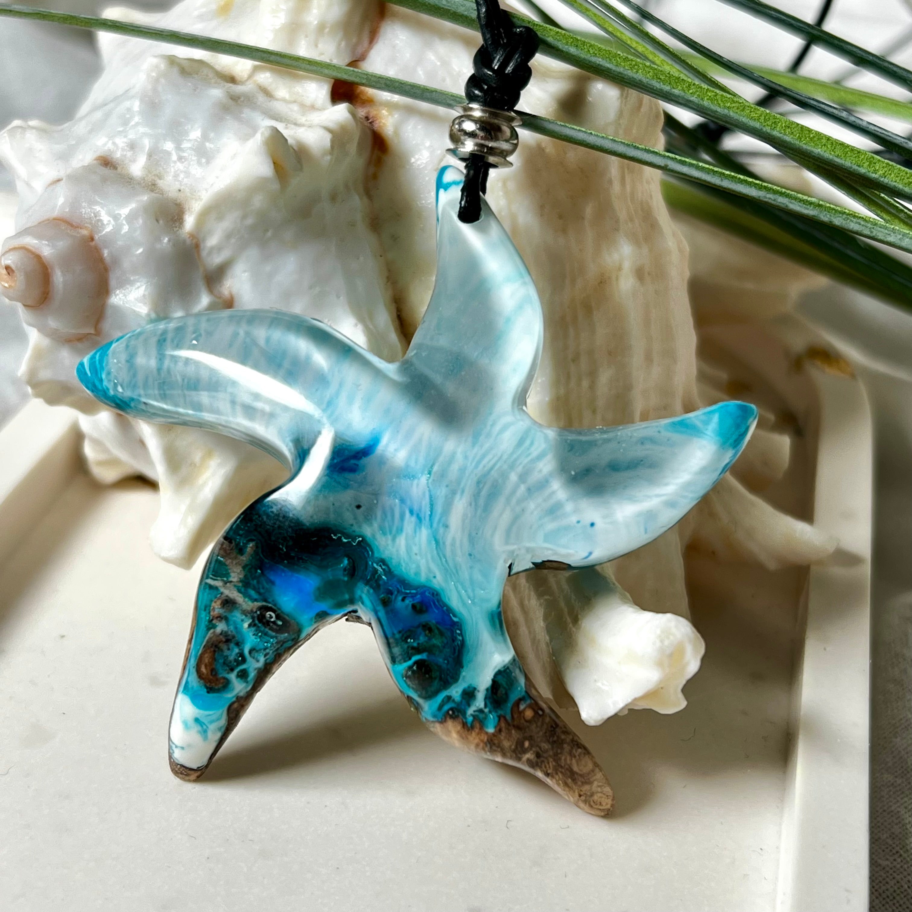 Handgefertigte Halskette in Seestern-Form mit Anhänger aus Naturholz und blau-weiß marmoriertem Epoxidharz. Das Schmuckstück ist einzigartig gestaltet und spiegelt maritimen Stil und Naturverbundenheit wider. Getragen an einem verstellbaren Lederband mit Holzperlen. Ideal für Meeresliebhaber und Naturfreunde.