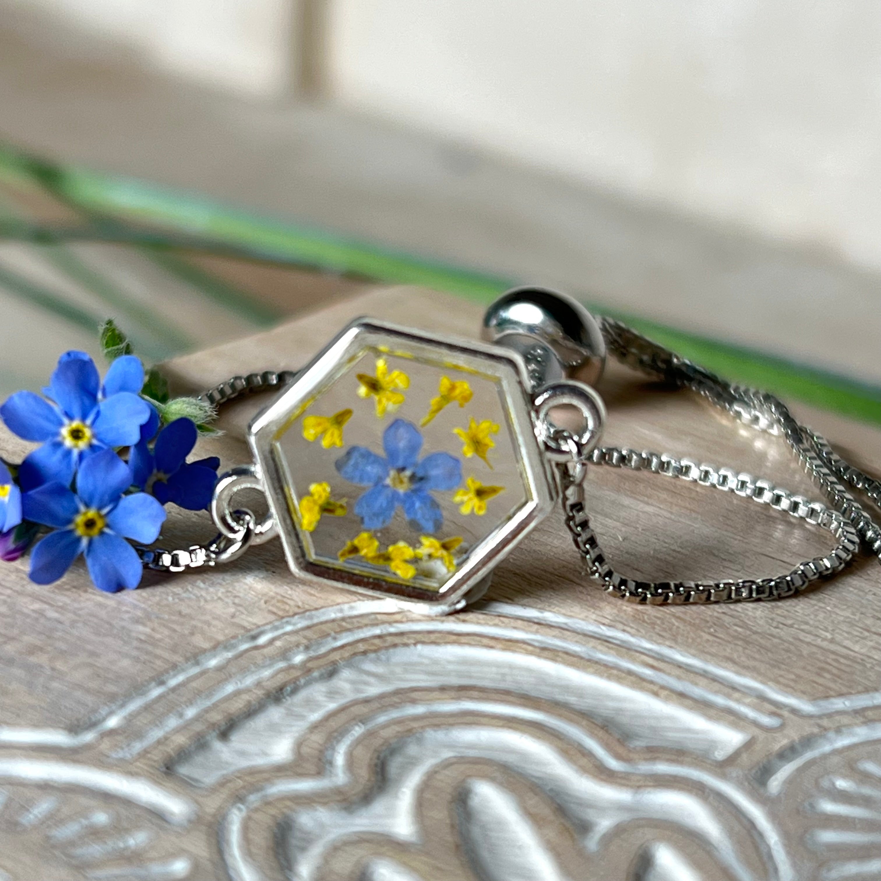 „Detailaufnahme eines handgefertigten Armbands mit echter blauer Vergissmeinnicht und feinen gelben Blüten in Harz, eingefasst in sechseckiger versilberter Lünette. Verstellbares Edelstahlarmband mit funkelnden Zirkonia-Endperlen – liebevolles Natur-Schmuckstück, ideal als Geschenk für Frauen.“