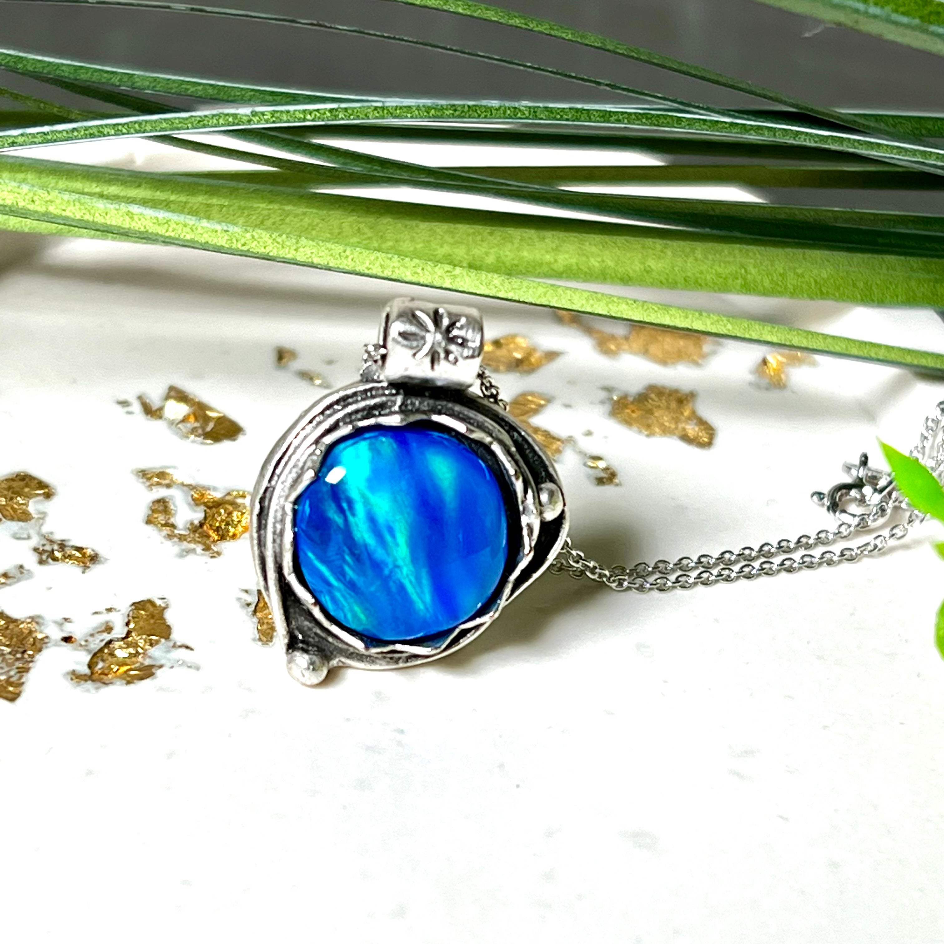 Handgemachter Anhänger mit rundem Cabochon aus Epoxidharz und schimmerndem synthetischem Opal. Die Farben changieren je nach Lichteinfall in Blau, Türkis und Grün. Der Opal sitzt in einer antik versilberten Fassung aus Messing (blei- & nickelfrei). Geliefert mit einer rhodinierten 925 Silberkette (45 cm). Jedes Stück ein Einzelstück – perfekt als stilvolles Geschenk oder persönlicher Glücksbringer.