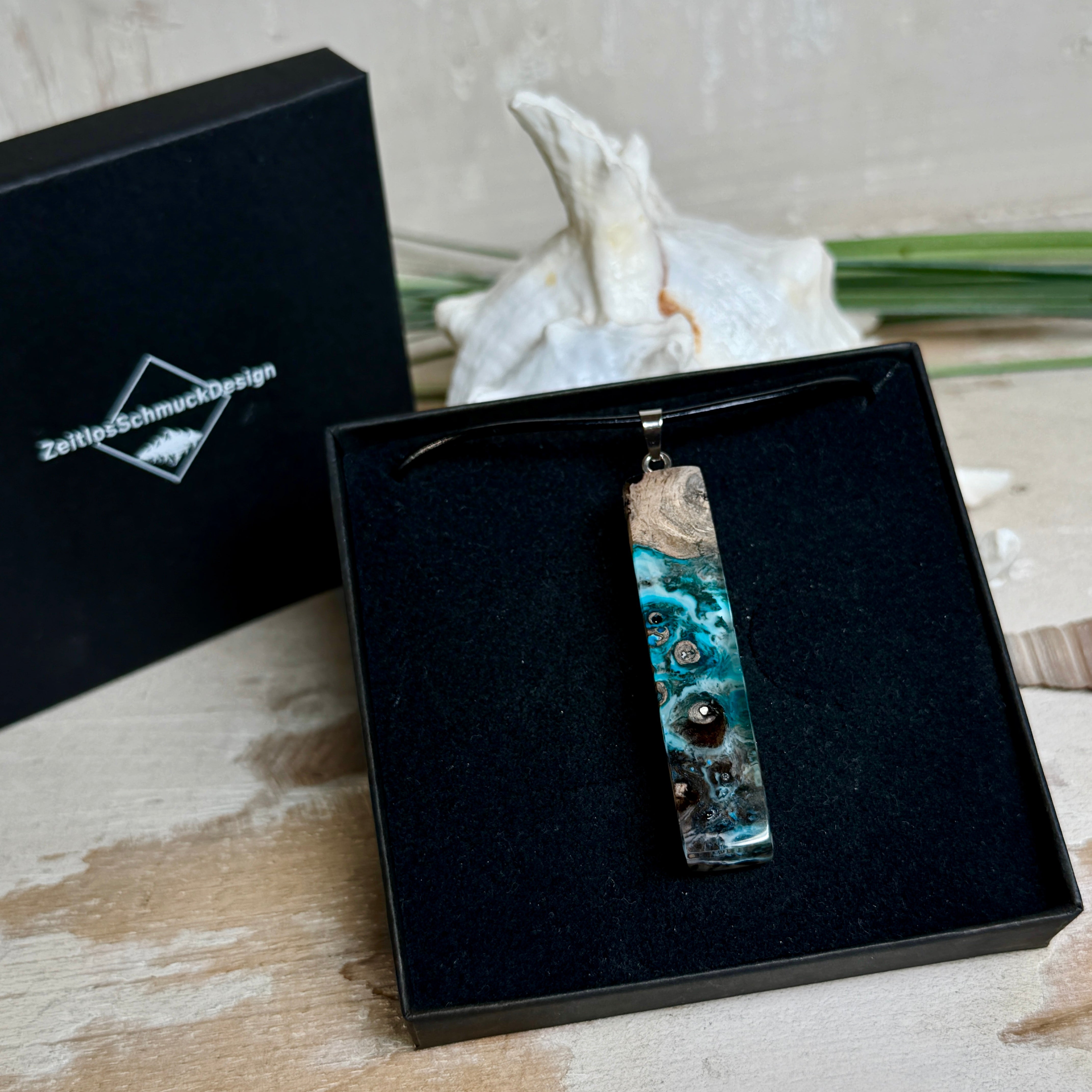 Rechteckiger Stabanhänger aus Maserholz und glasklarem Epoxidharz in tiefen Blau- und Türkistönen mit organischer Lagunen/Riff-Optik. Hochglanzpoliert, ca. 5,3 × 1,2 cm. Der Anhänger hängt an einer schwarzen Lederkette mit Edelstahl-Karabiner und 5-cm-Verlängerung (45 + 5 cm); die Anhängerschlaufe ist ebenfalls aus Edelstahl. Handgefertigtes Unikat, Lieferung in edler Schmuckbox – maritimer Look.