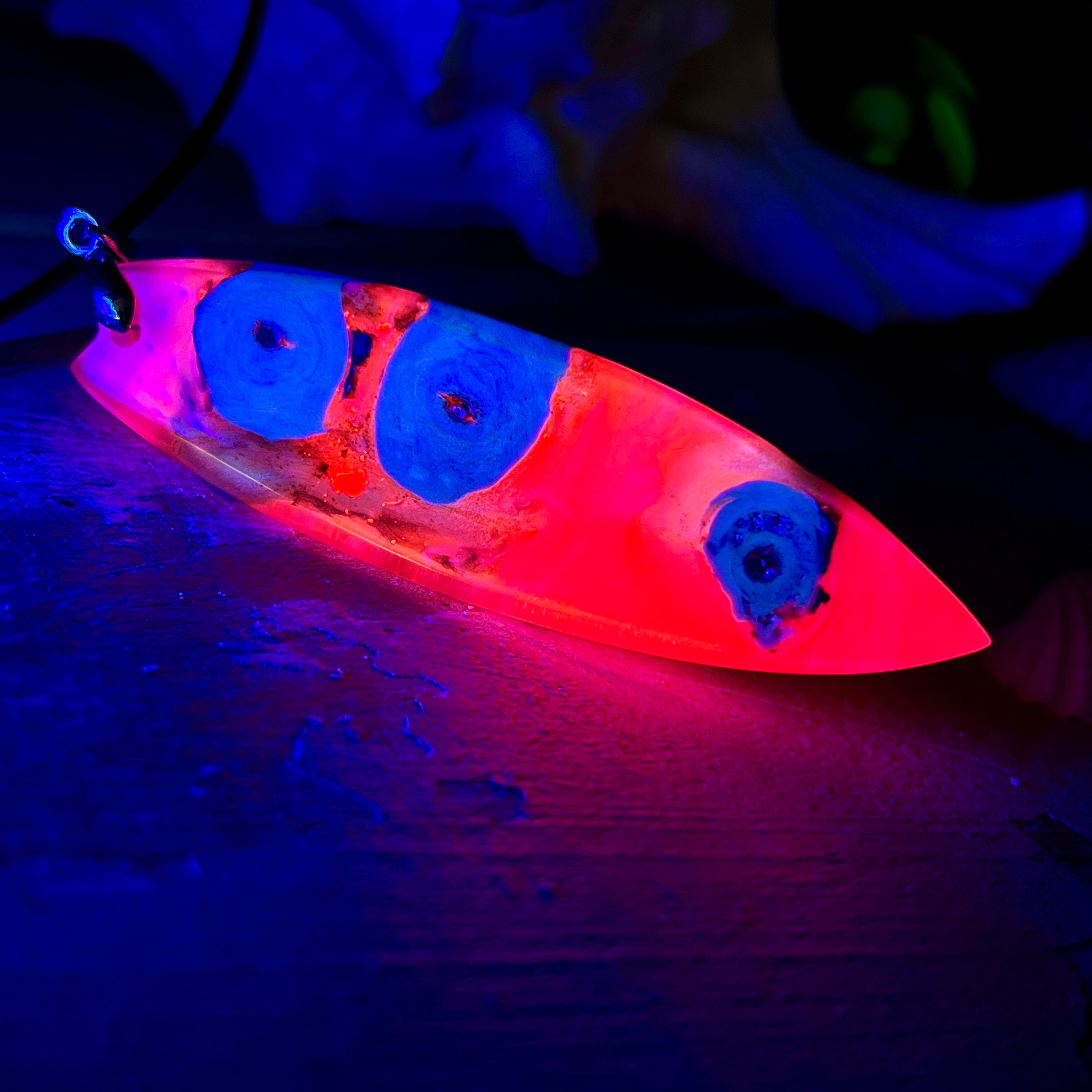 Handgefertigter Surfbrett-Anhänger „Flame Surf“ aus Maserholz und fluoreszierendem Harz in kräftigem Pink. Das Schmuckstück leuchtet bei Schwarzlicht und wird an einer schwarzen Lederkette mit Edelstahl-Karabiner (45 cm + 5 cm Verlängerung) geliefert. Größe ca. 6,7 × 2 cm. Ein Unikat für Surf- und Strandliebhaber, edel verarbeitet und mit Liebe gefertigt.