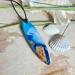 Handgefertigter Surfbrett-Anhänger „Ocean Glide“ aus Maserholz und Epoxidharz in verschiedenen Blau-, und Weißtönen. Der ca. 6,5 × 2 cm große Anhänger wird an einem verstellbaren Lederband (26–46 cm) mit Holzperlen geliefert. Ein maritimes Unikat mit natürlicher Holzmaserung, das die Schönheit des Meeres einfängt.