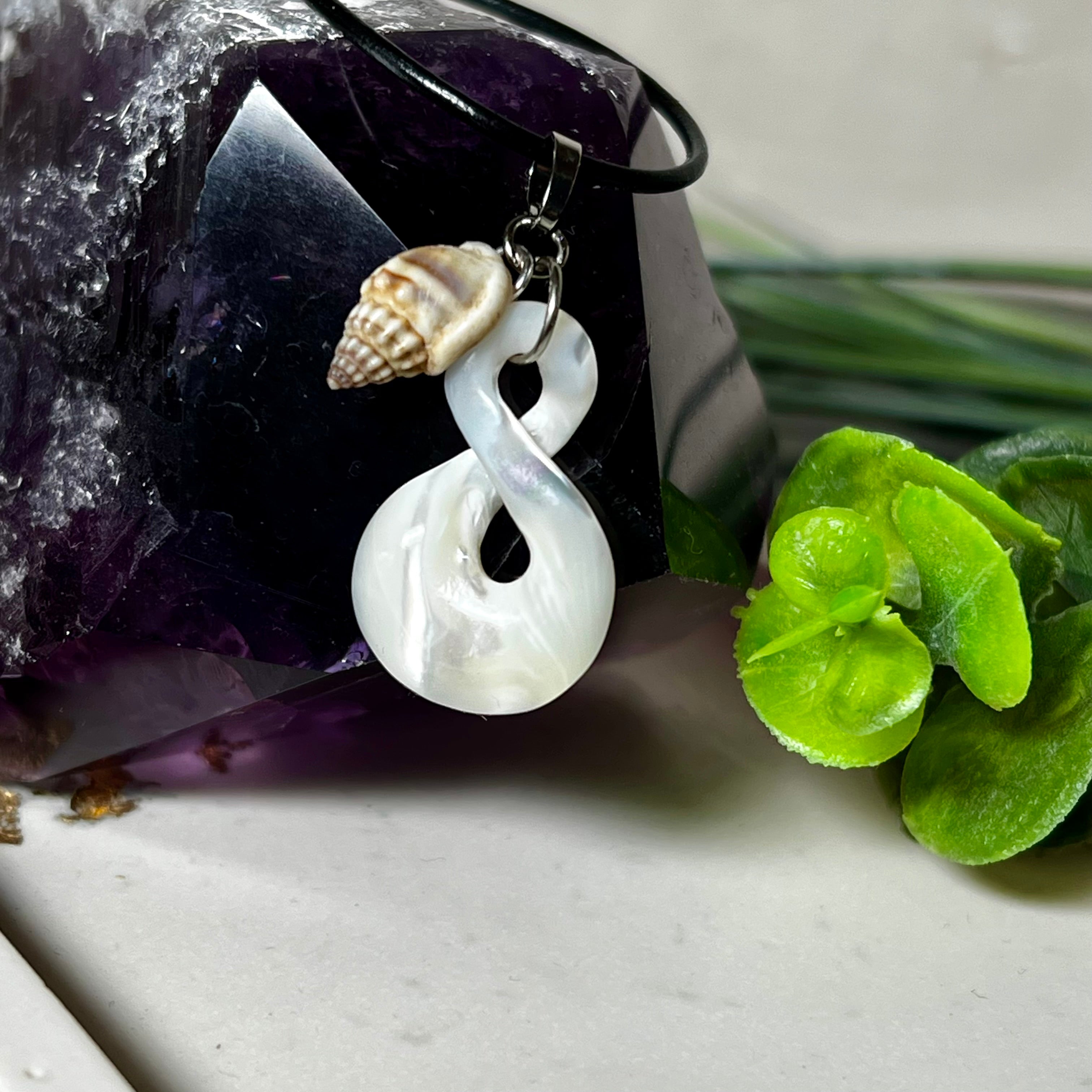 Detailaufnahme eines handgefertigten Anhängers aus echtem Perlmutt in Schlaufenform, kombiniert mit einer kleinen echten Muschel als Charm-Anhänger. Der Schmuck hängt an einer feinen schwarzen Lederschnur mit Edelstahlverschluss und symbolisiert Harmonie und Unendlichkeit. Ideal für Meeresliebhaberinnen und Fans von Naturmaterialien.