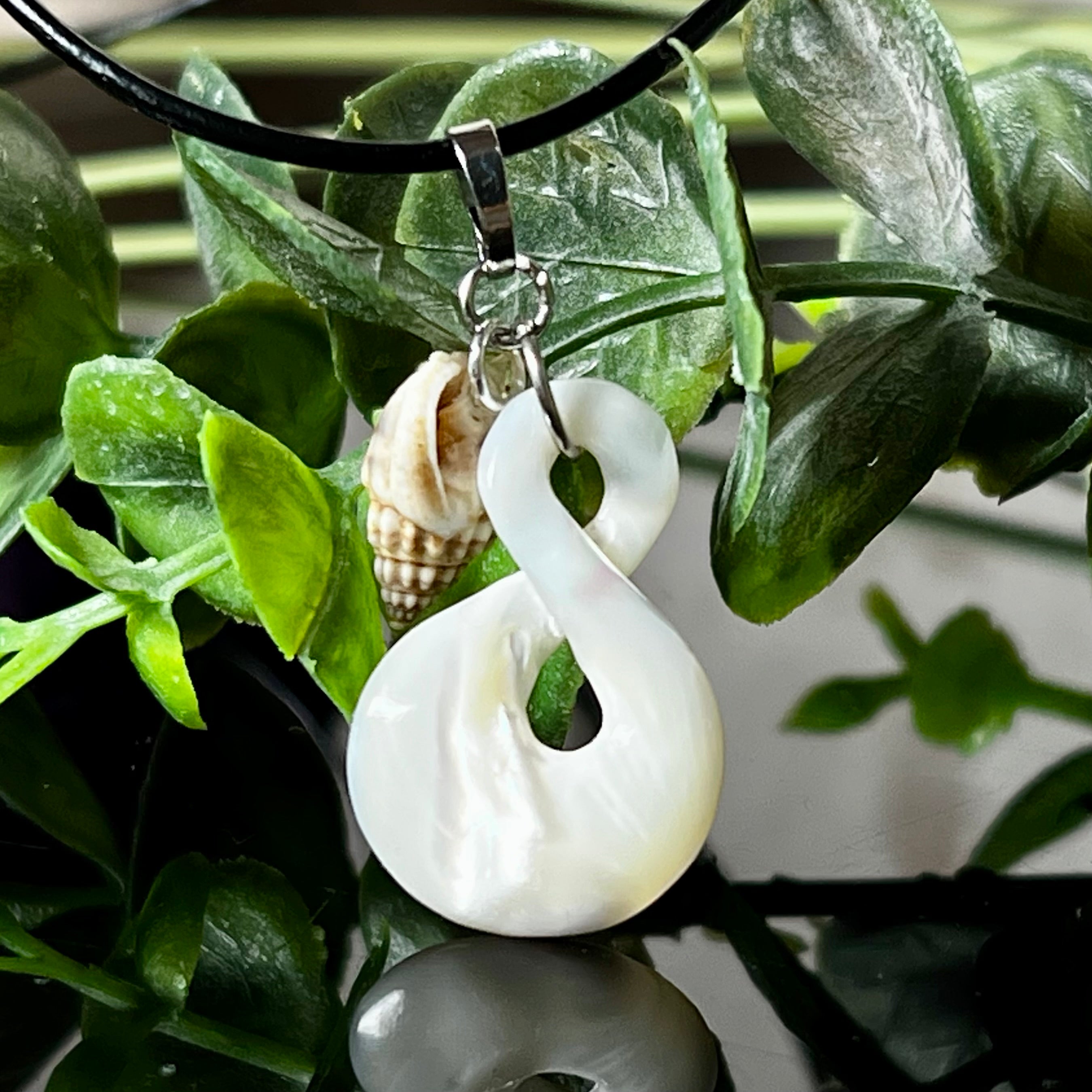 Detailaufnahme eines handgefertigten Anhängers aus echtem Perlmutt in Schlaufenform, kombiniert mit einer kleinen echten Muschel als Charm-Anhänger. Der Schmuck hängt an einer feinen schwarzen Lederschnur mit Edelstahlverschluss und symbolisiert Harmonie und Unendlichkeit. Ideal für Meeresliebhaberinnen und Fans von Naturmaterialien.