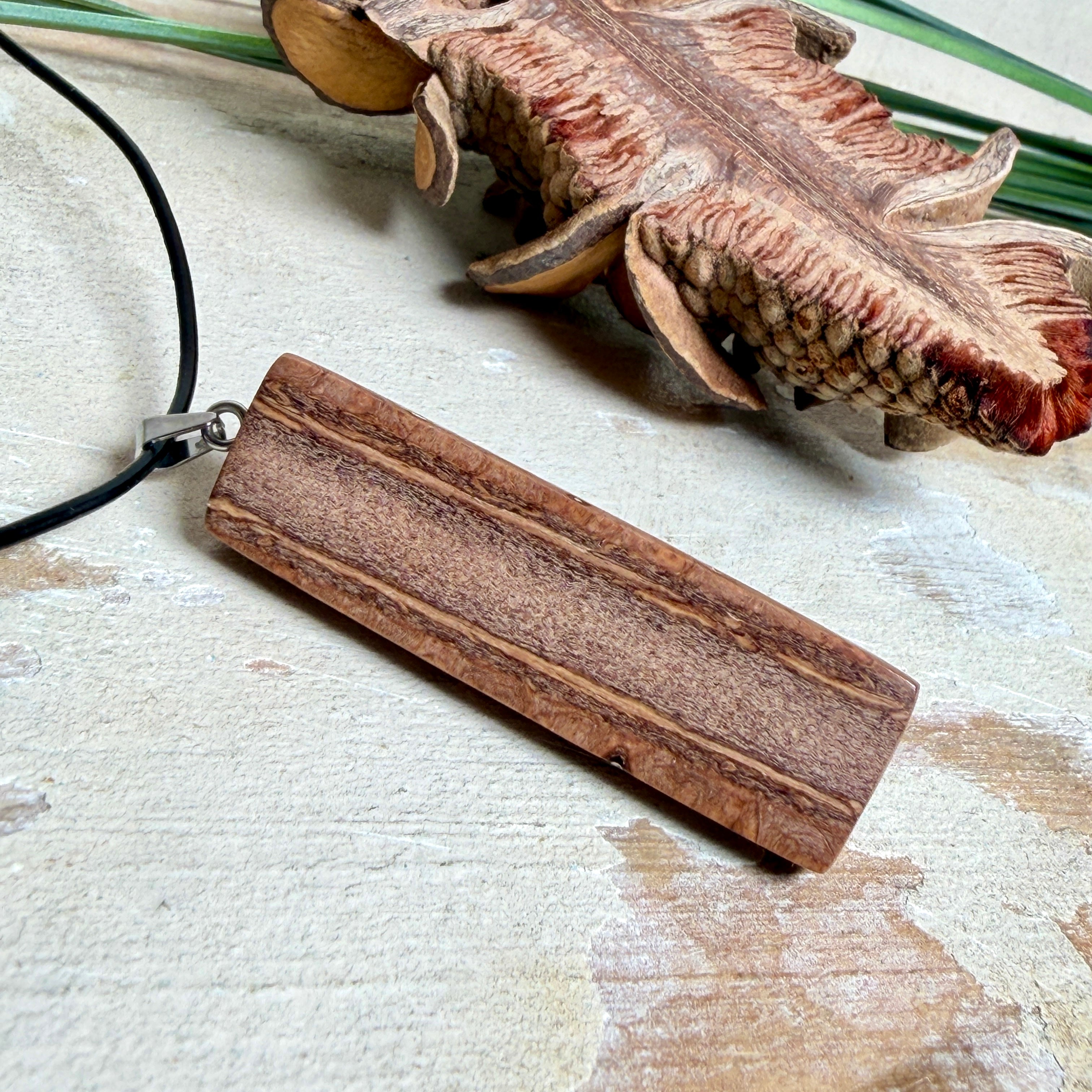 Handgefertigter Halsanhänger aus echtem Banksiazapfen, veredelt mit rotem Harz in den natürlichen Kammern; ca. 5,5 × 1,6 cm. Mit Anhängerschlaufe aus Edelstahl an schwarzer Lederkette (45 cm + 5 cm Verlängerung) mit Edelstahl-Karabiner. Detailaufnahme bzw. Tragebild vor neutralem, natürlichem Hintergrund.