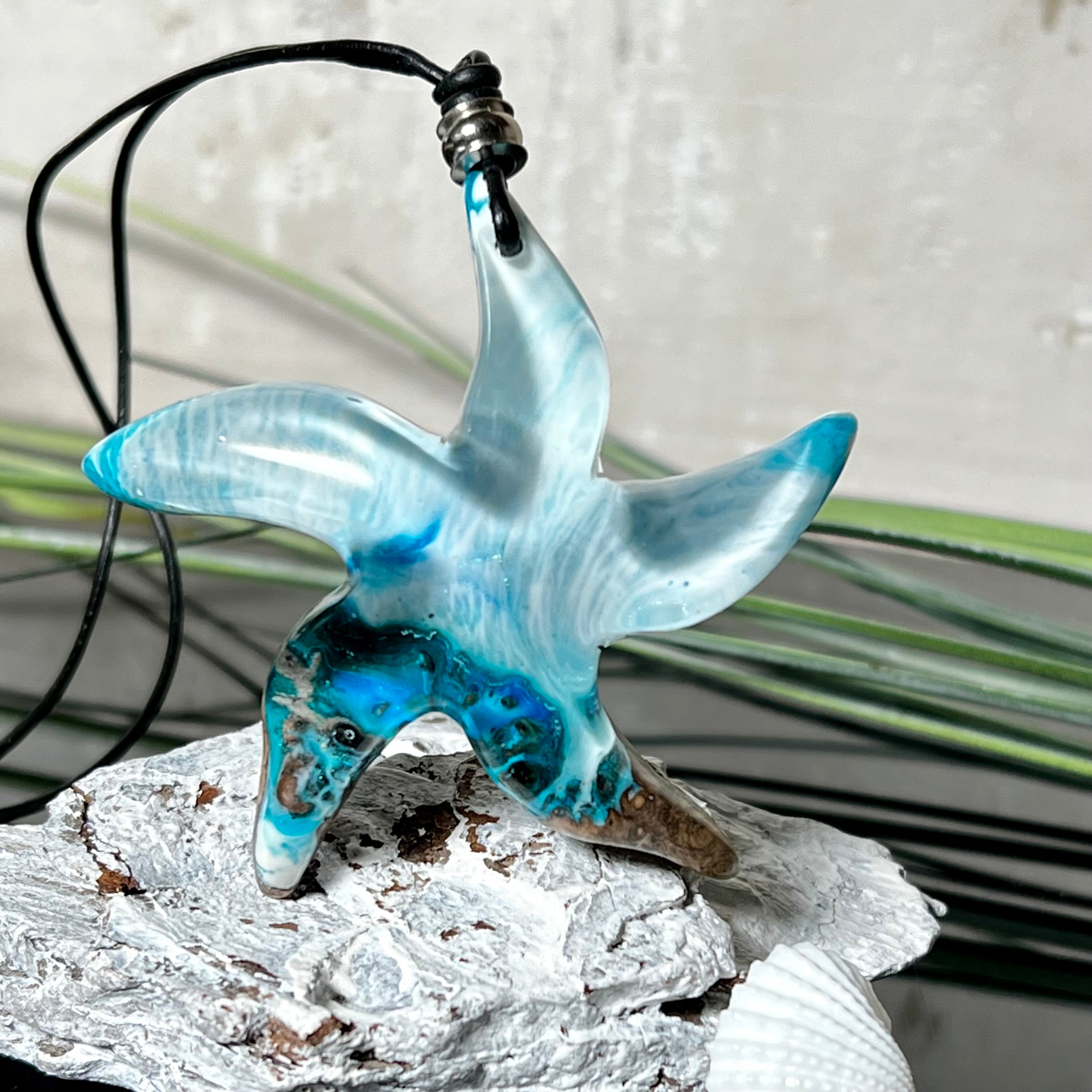 Handgefertigte Halskette in Seestern-Form mit Anhänger aus Naturholz und blau-weiß marmoriertem Epoxidharz. Das Schmuckstück ist einzigartig gestaltet und spiegelt maritimen Stil und Naturverbundenheit wider. Getragen an einem verstellbaren Lederband mit Holzperlen. Ideal für Meeresliebhaber und Naturfreunde.