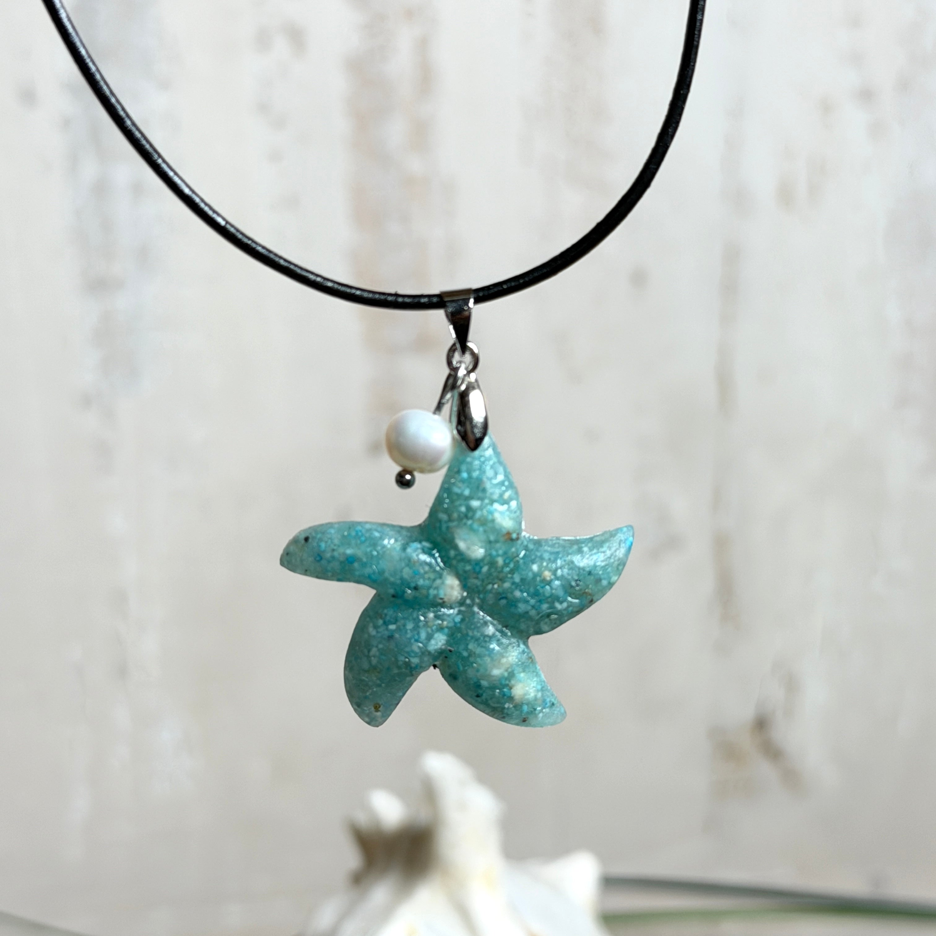 Seestern-Anhänger aus transparentem Harz mit echtem Meersand ( leicht eingefärbt), in blautürkis Strand-Look, mit kleiner Süßwasserperle als Anhänger-Detail. Edelstahlöse, schwarze Lederkette mit Edelstahl-Karabiner und Verlängerung (45 cm + 5 cm). Anhänger ca. 3,5 × 3,5 cm. Handgefertigtes Unikat; vereinzelt kleine Luftbläschen möglich, typisch für Handarbeit.