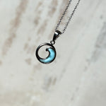 Feine Wellen-Kette aus rhodiniertem 925 Silber. Der runde Anhänger zeigt eine stilisierte Welle mit hellblauem Larimar-Cabochon, eingefasst in poliertem Silber. Größe des Anhängers ca. 23 x 13 mm, an einer 45 cm langen, 1 mm starken, rhodinierten Silberkette. 