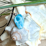 Larimar Freeform-Anhänger auf schwarzer Lederkette. Beidseitig tragbarer Trommelstein, ca. 2 x 1,8 cm groß, mit Silberöse. Der seltene, kostbare Larimar zeigt lebendige Blau- und Weißnuancen wie karibisches Lagunenwasser. Lederkette 45 cm mit Edelstahl-Karabiner und 5 cm Edelstahl-Verlängerung, fotografiert vor einer großen Muschel im maritimen Stil.