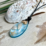 Handgefertigter Tropfenanhänger aus Maserholz und klarem Epoxidharz in Aquatönen. Im Harz sind echte Muscheln und ein kleiner Seestern eingebettet. Durch die besondere Mehrschicht-Gießtechnik wirkt es, als würden die Muscheln im Wasser schweben. Größe ca. 3,8 × 2 cm. Verstellbares Lederband (24–44 cm) mit Holzperlen. Maritimes Unikat.