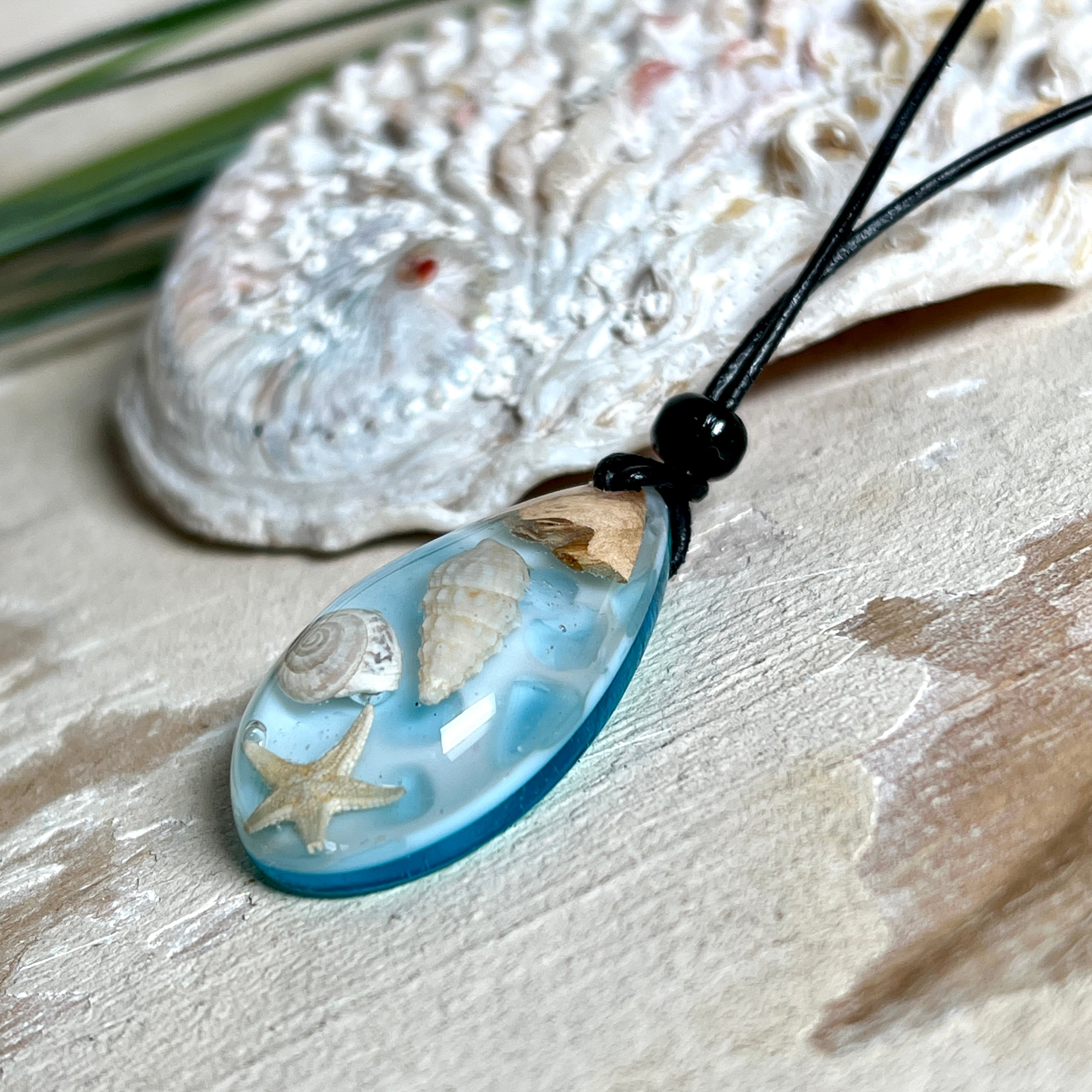 Handgefertigter Tropfenanhänger aus Maserholz und klarem Epoxidharz in Aquatönen. Im Harz sind echte Muscheln und ein kleiner Seestern eingebettet. Durch die besondere Mehrschicht-Gießtechnik wirkt es, als würden die Muscheln im Wasser schweben. Größe ca. 3,8 × 2 cm. Verstellbares Lederband (24–44 cm) mit Holzperlen. Maritimes Unikat.