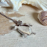 Feine Kette aus rhodiniertem 925 Silber mit Wasserschildkröten-Anhänger. Der ca. 25 x 18 mm große Anhänger zeigt eine Schildkröte, deren Panzer aus mehreren hellblauen Larimar-Inlays besteht. Seltener, kostbarer Larimar mit typischen wolkigen Strukturen, an einer ca. 45 cm langen, 1 mm starken, auf Platin rhodinierten Silberkette. Fotografiert in schwarzer Schmuckbox und vor maritimer Dekoration mit Muscheln.