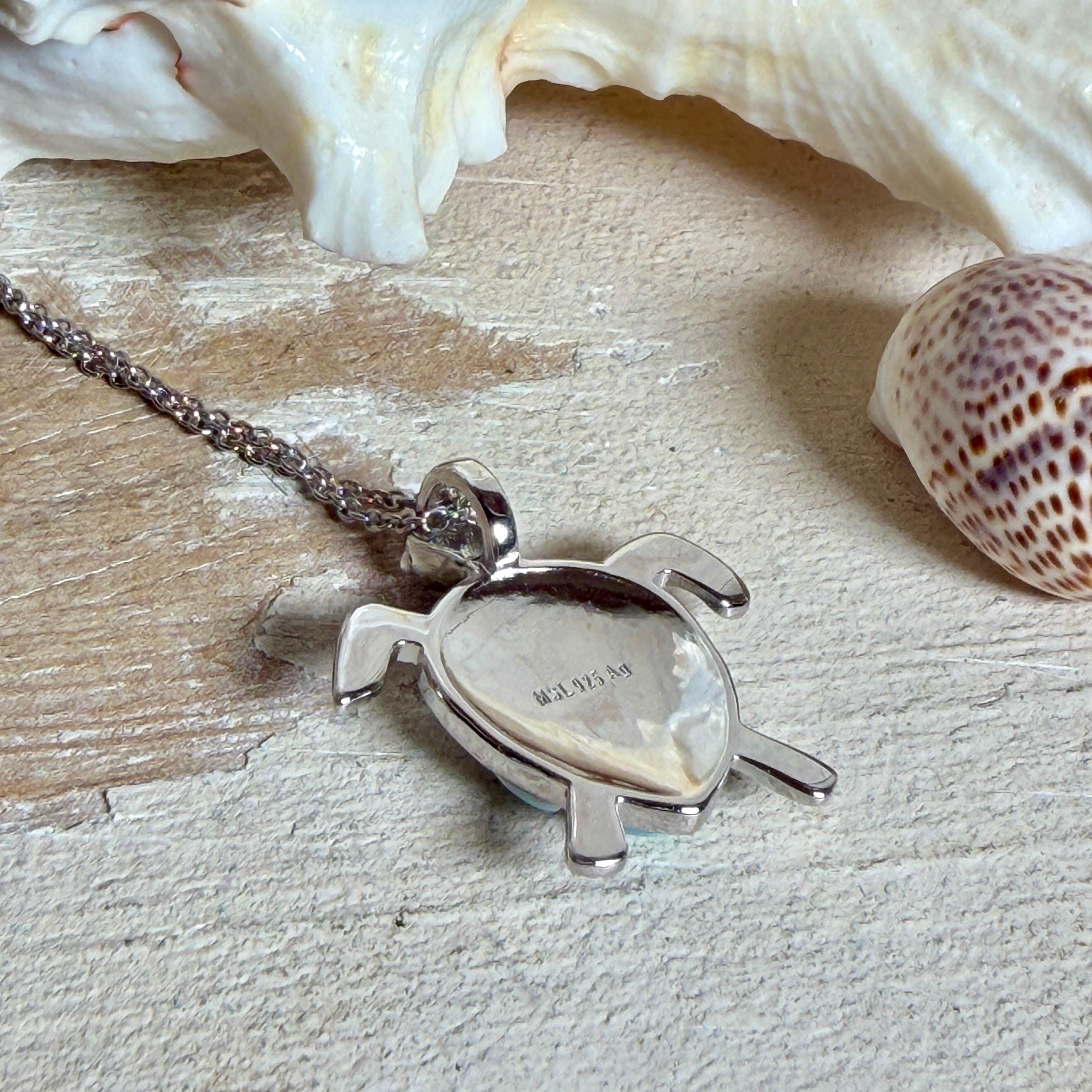 Feine Kette aus rhodiniertem 925 Silber mit Wasserschildkröten-Anhänger. Der ca. 25 x 18 mm große Anhänger zeigt eine Schildkröte, deren Panzer aus mehreren hellblauen Larimar-Inlays besteht. Seltener, kostbarer Larimar mit typischen wolkigen Strukturen, an einer ca. 45 cm langen, 1 mm starken, auf Platin rhodinierten Silberkette. Fotografiert in schwarzer Schmuckbox und vor maritimer Dekoration mit Muscheln.